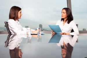 Retail pharmacy manager talks to a new job applicant. They are both women, and they both wear white shirts. We can see a panorama of a city behind the windows. 