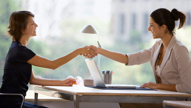 Two women, one in blue and one in cream dress, shake hands in an interview